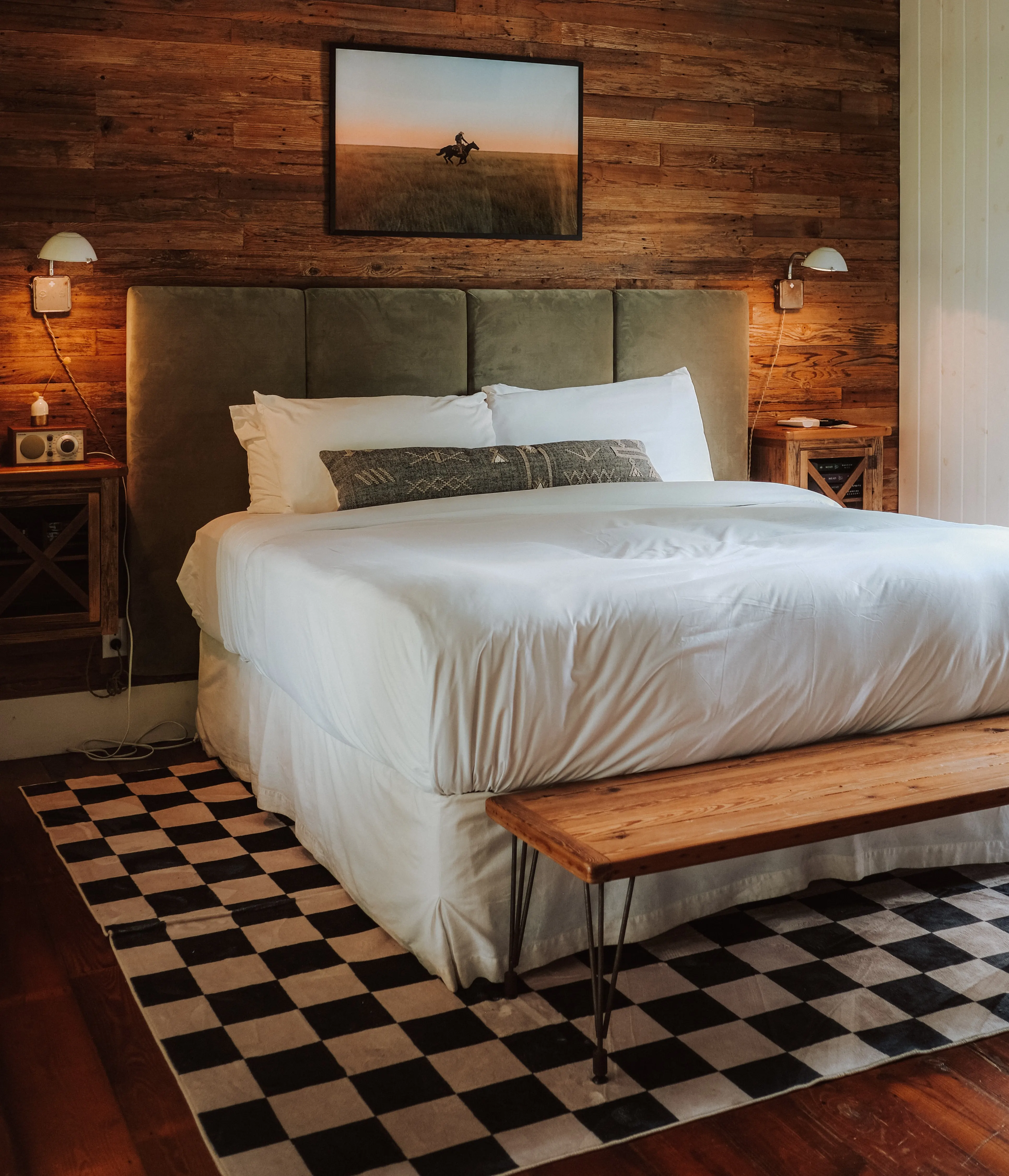 King bed against a reclaimed-barnwood wall under a teal ceiling, checkerboard rug and pine bench, Camp Comfort