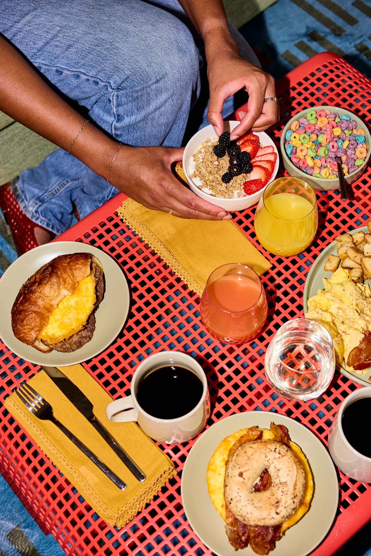 Breakfast spread on a red mesh table: croissant sandwich, granola bowl, cereal and coffee, La Piscine