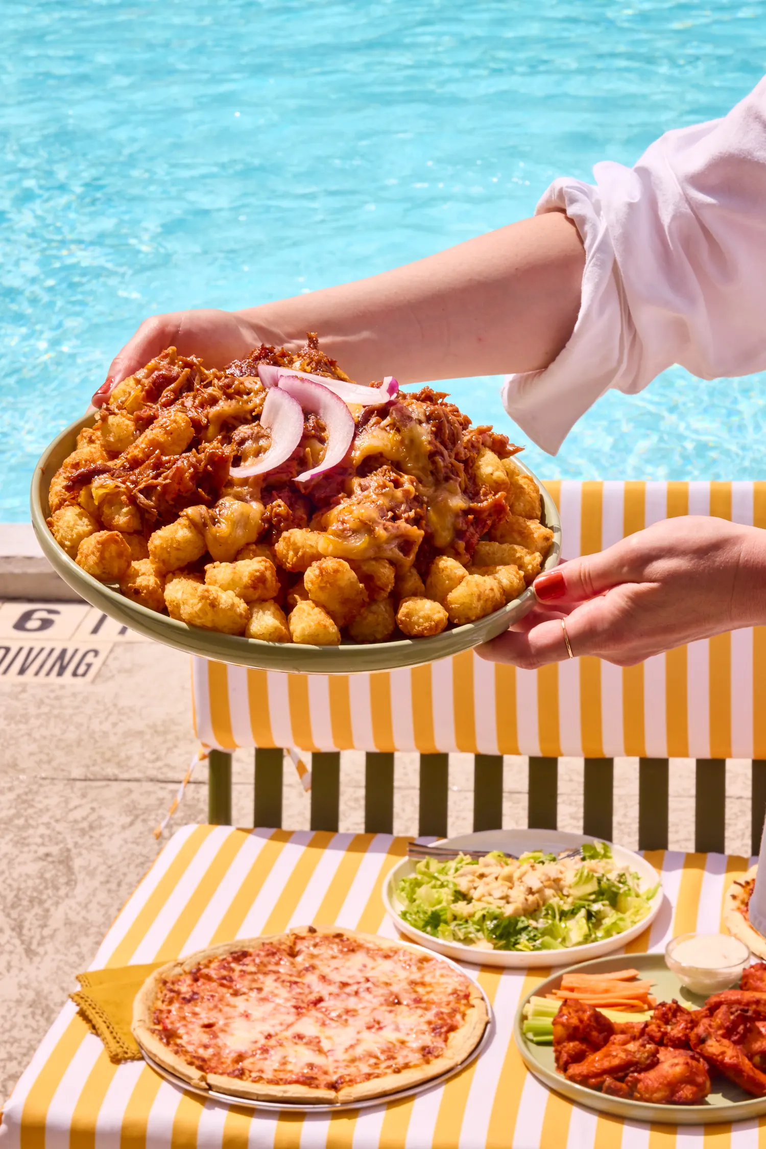 Loaded brisket tots held over a yellow striped table at the pool's edge, La Piscine