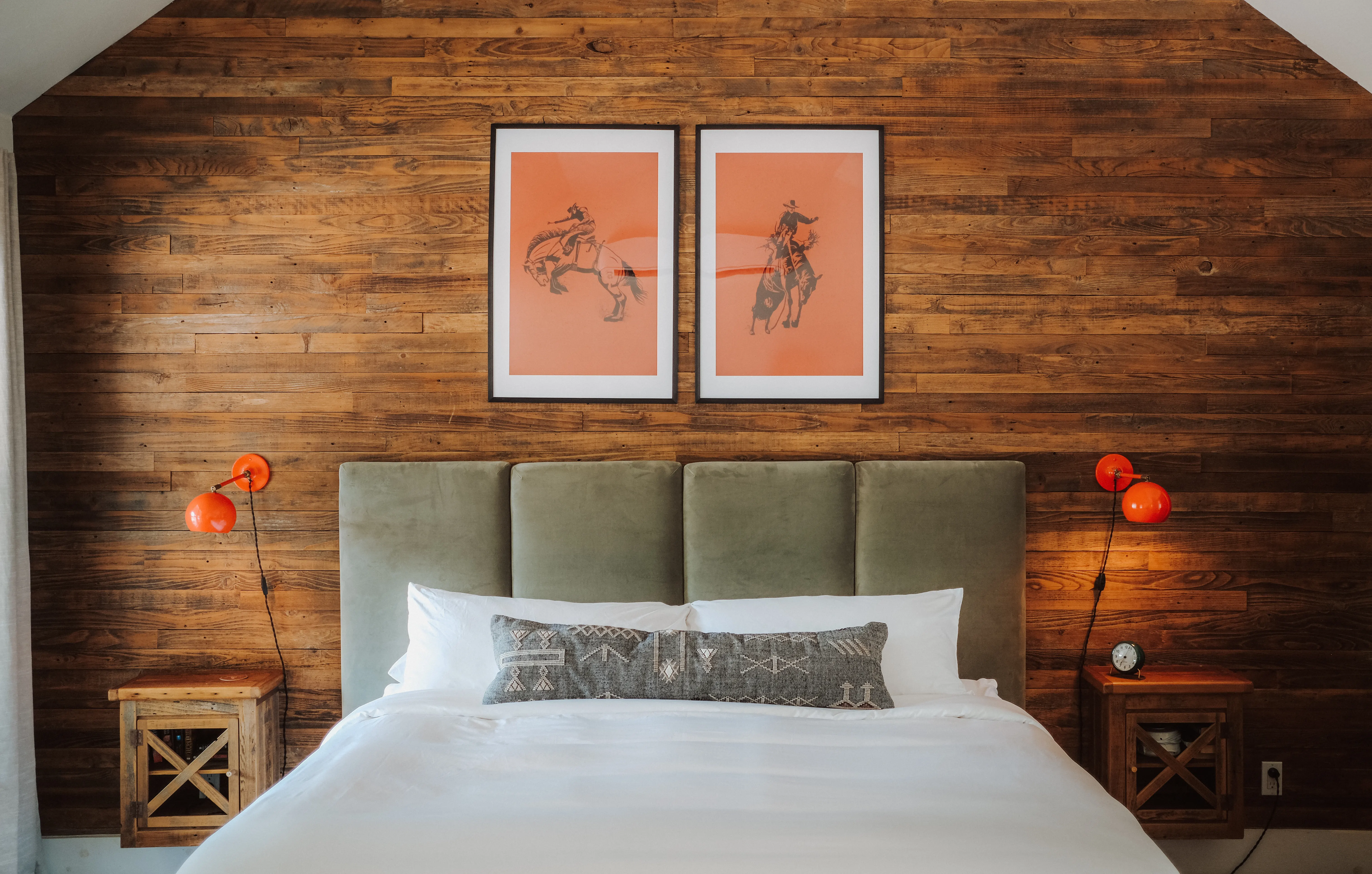King bed with a sage velvet headboard under two orange bronc-rider prints, orange sconces on reclaimed barnwood
