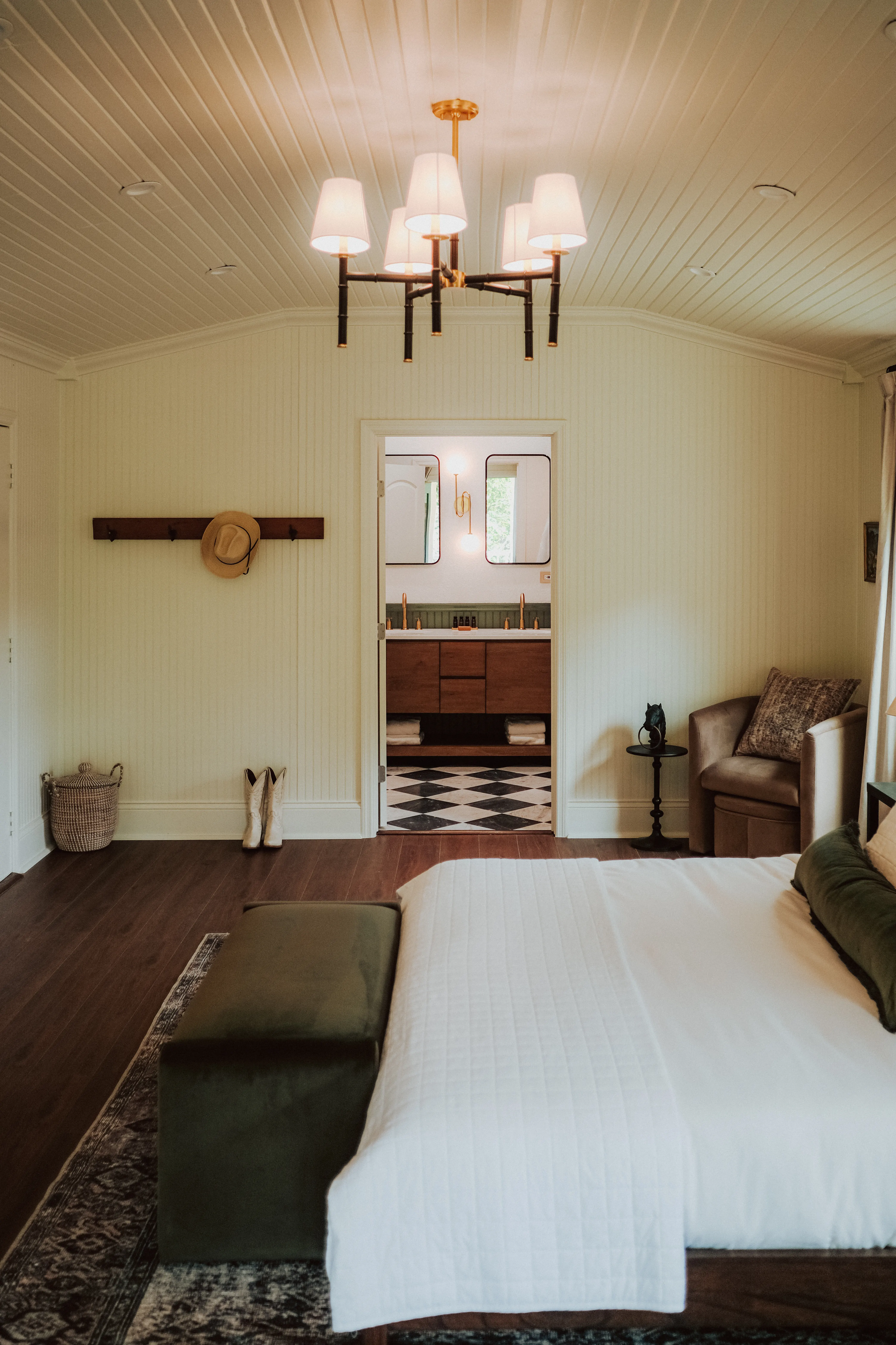 King room under a brass chandelier, a cowboy hat on the peg rail and a checkerboard-floored bath beyond