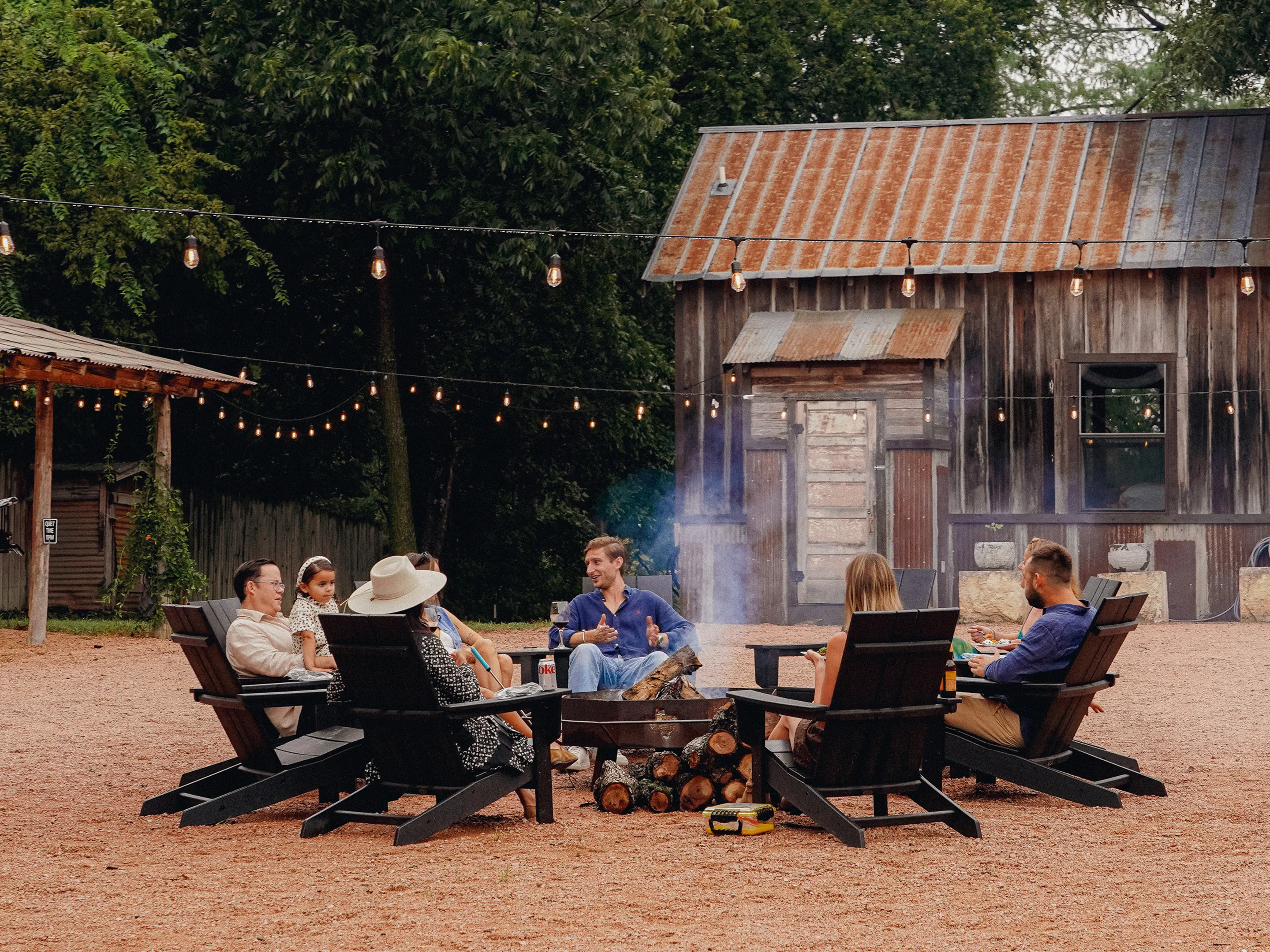 Guests around the fire pit under string lights at dusk, Camp Comfort