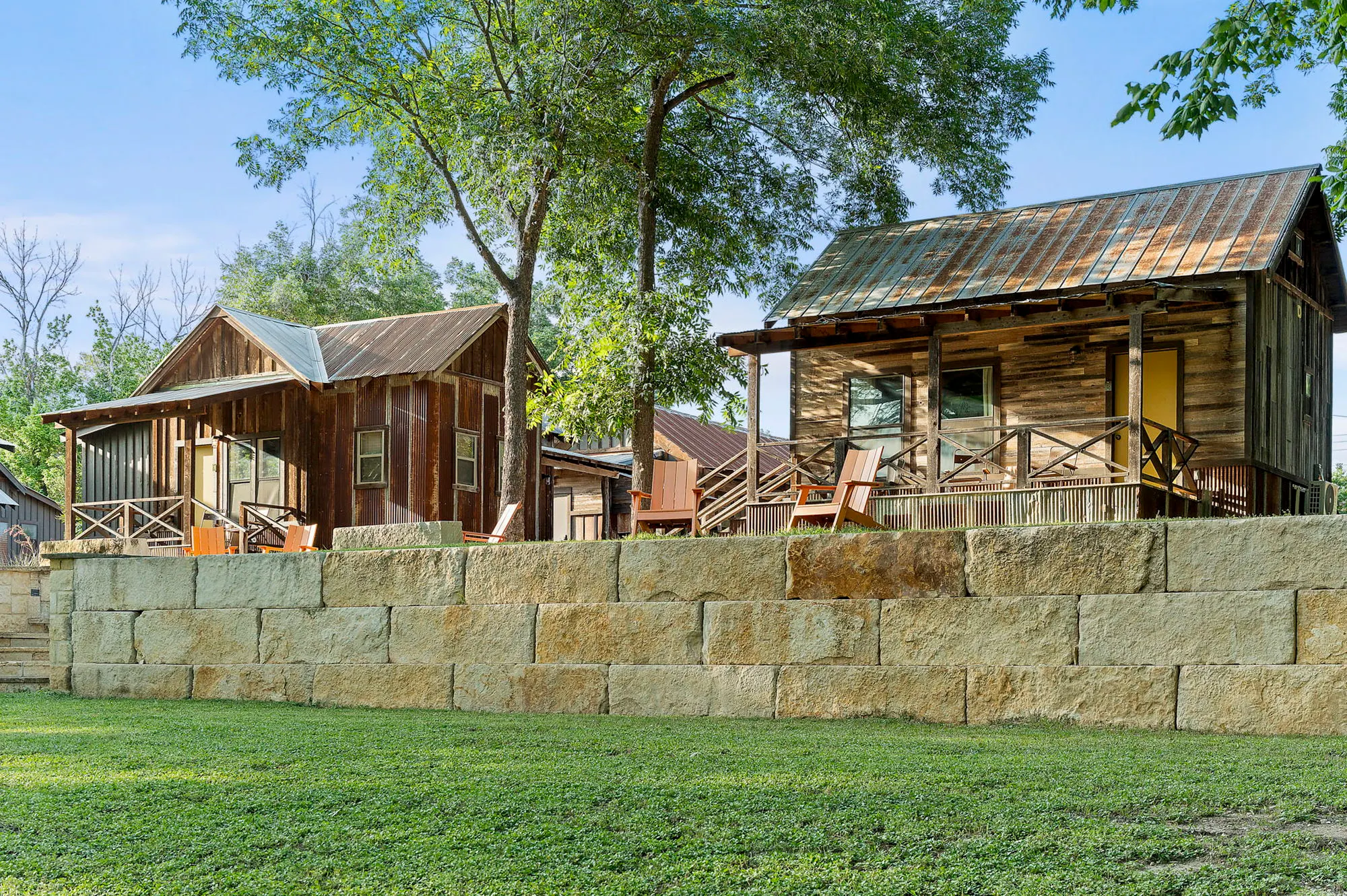 Reclaimed-wood cabins with tin roofs and orange chairs above a limestone wall and lawn