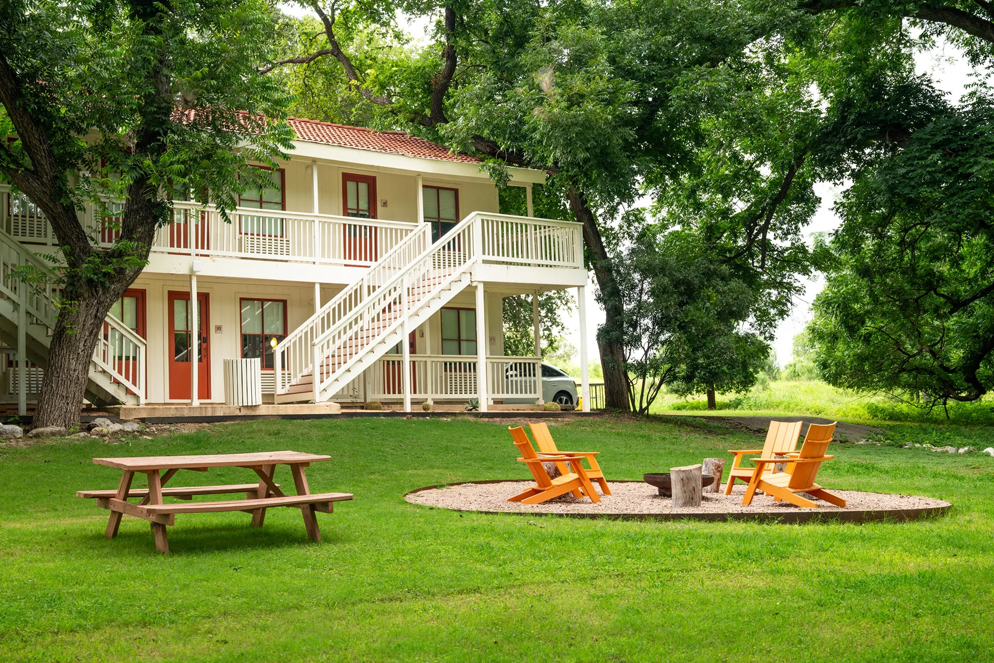 Two-storey lodge behind a lawn with a picnic table and orange chairs around a fire pit, Hacienda del Rio