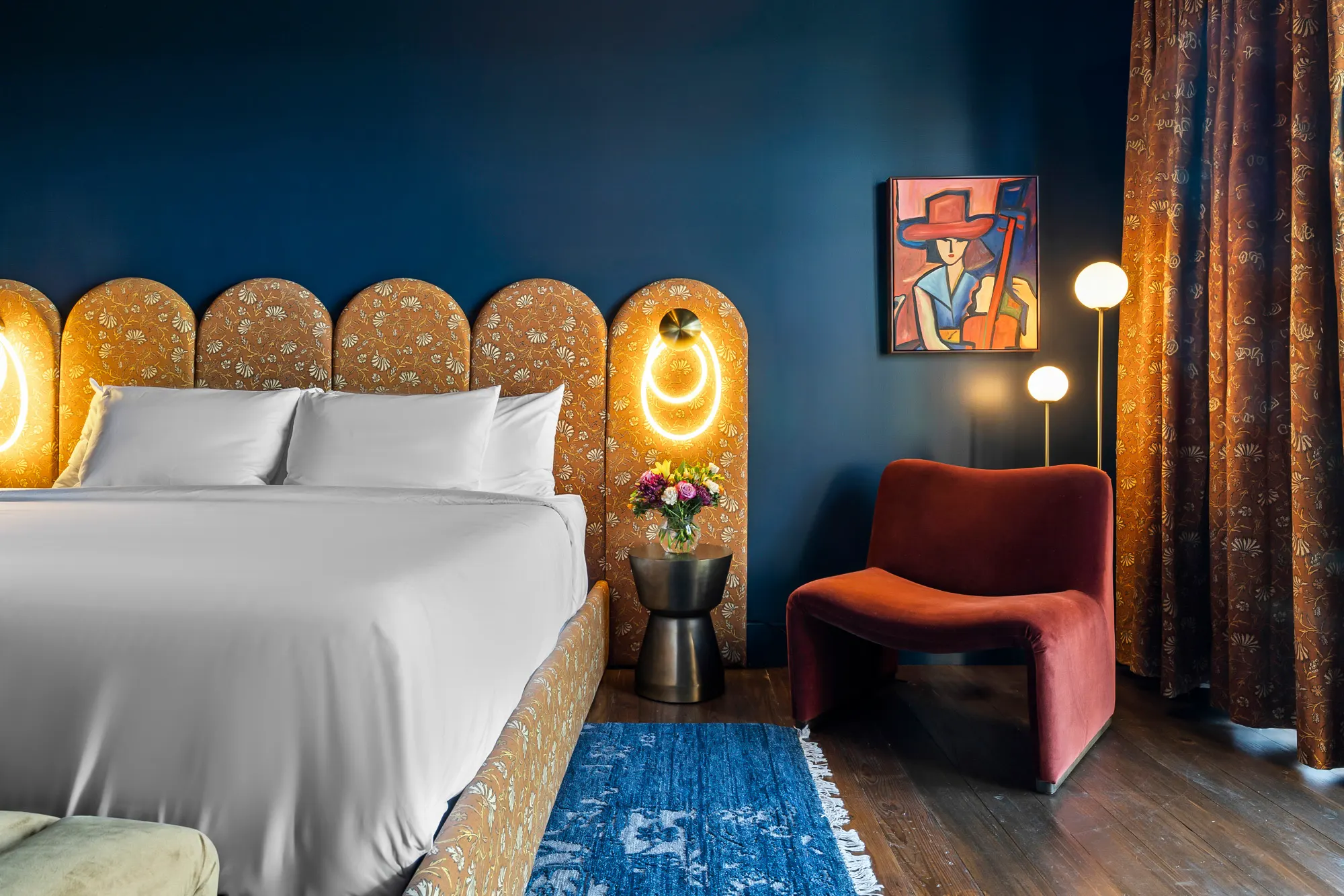 Navy king room with a scalloped floral headboard, ring sconces, a rust velvet chair and a blue rug, Gruene River Inn