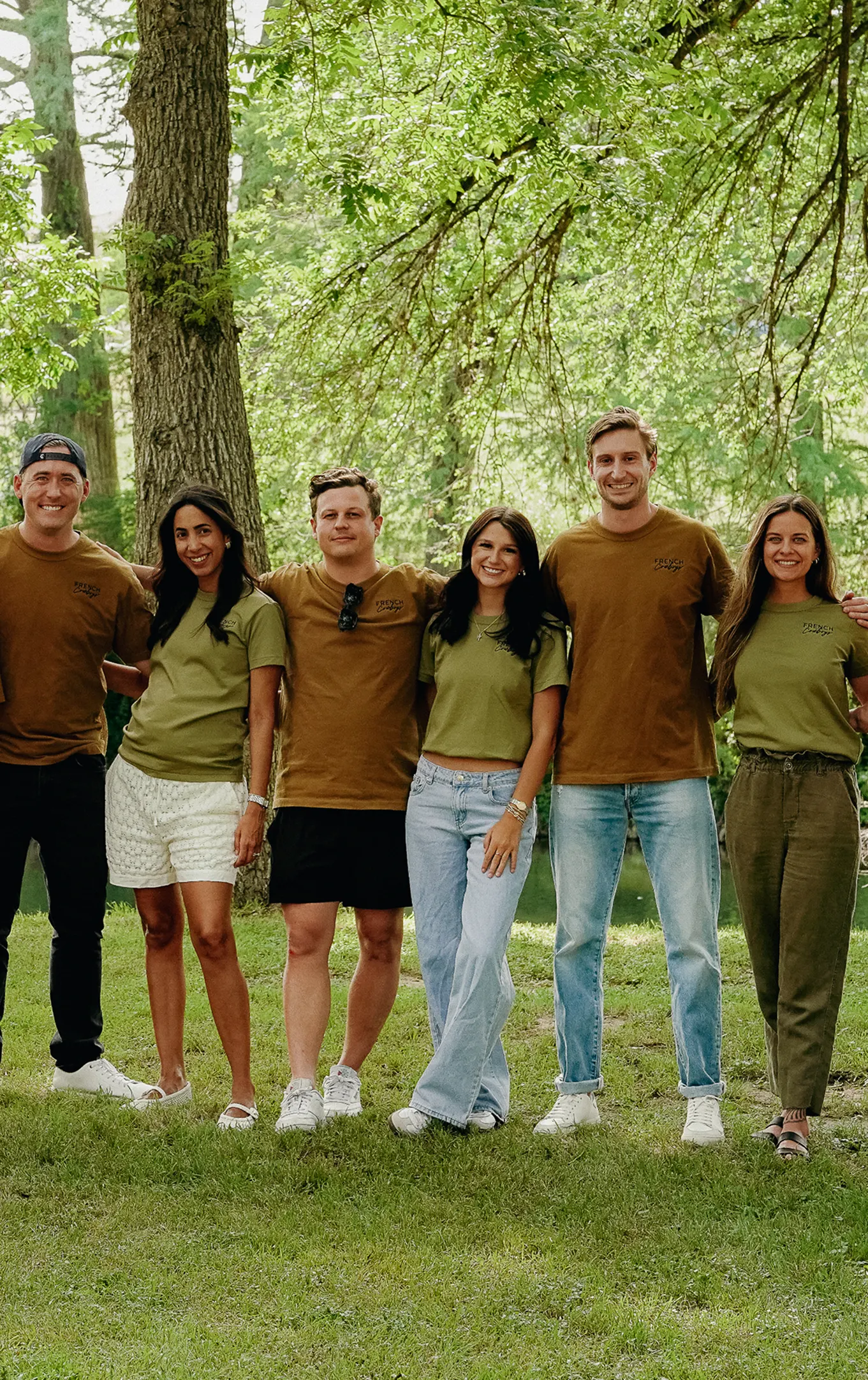 The French Cowboys team in matching olive and brown shirts, standing arm in arm on the grass by a river