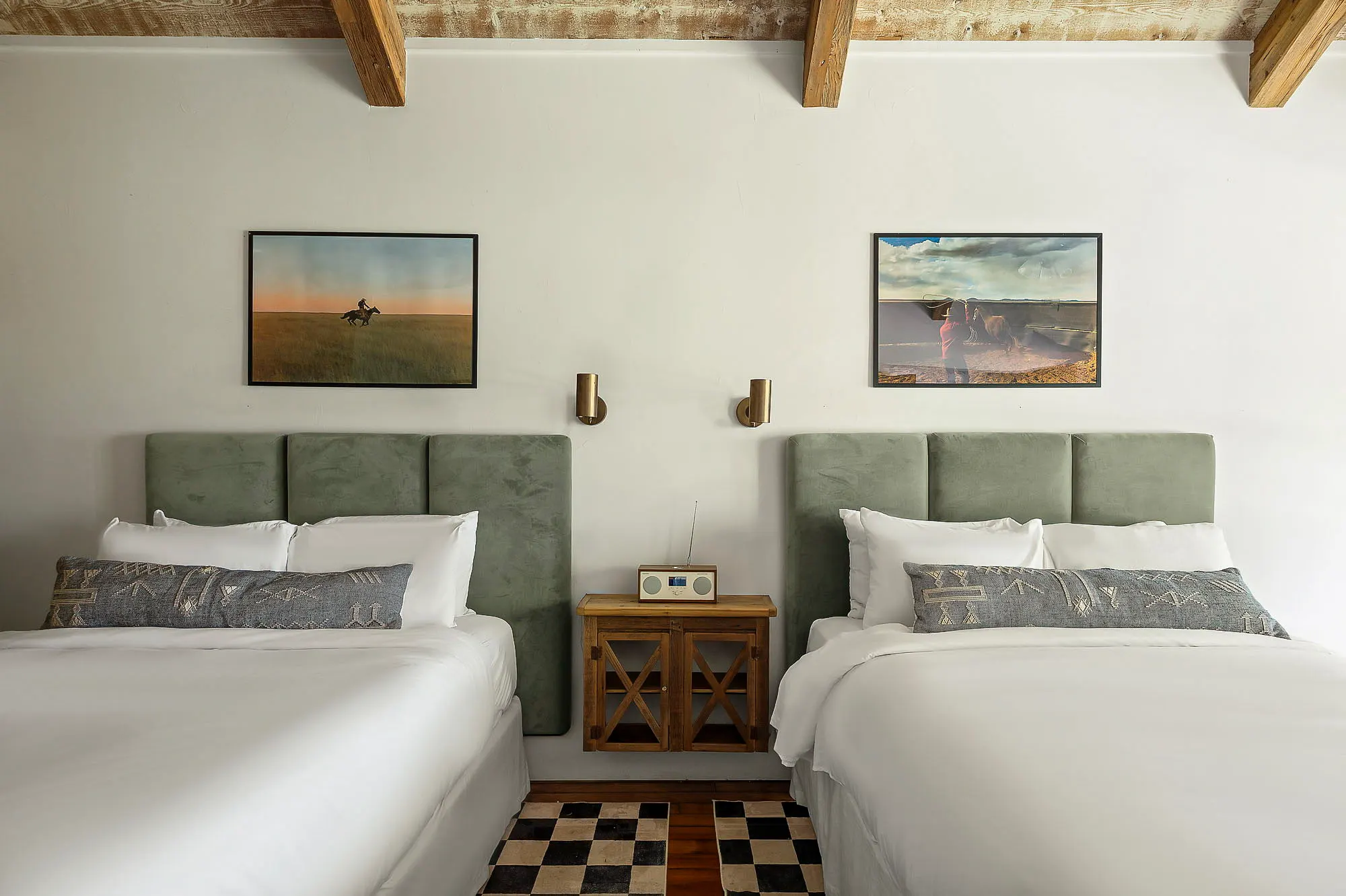 Two queen beds with sage velvet headboards under framed rider photographs and reclaimed beams, Camp Comfort