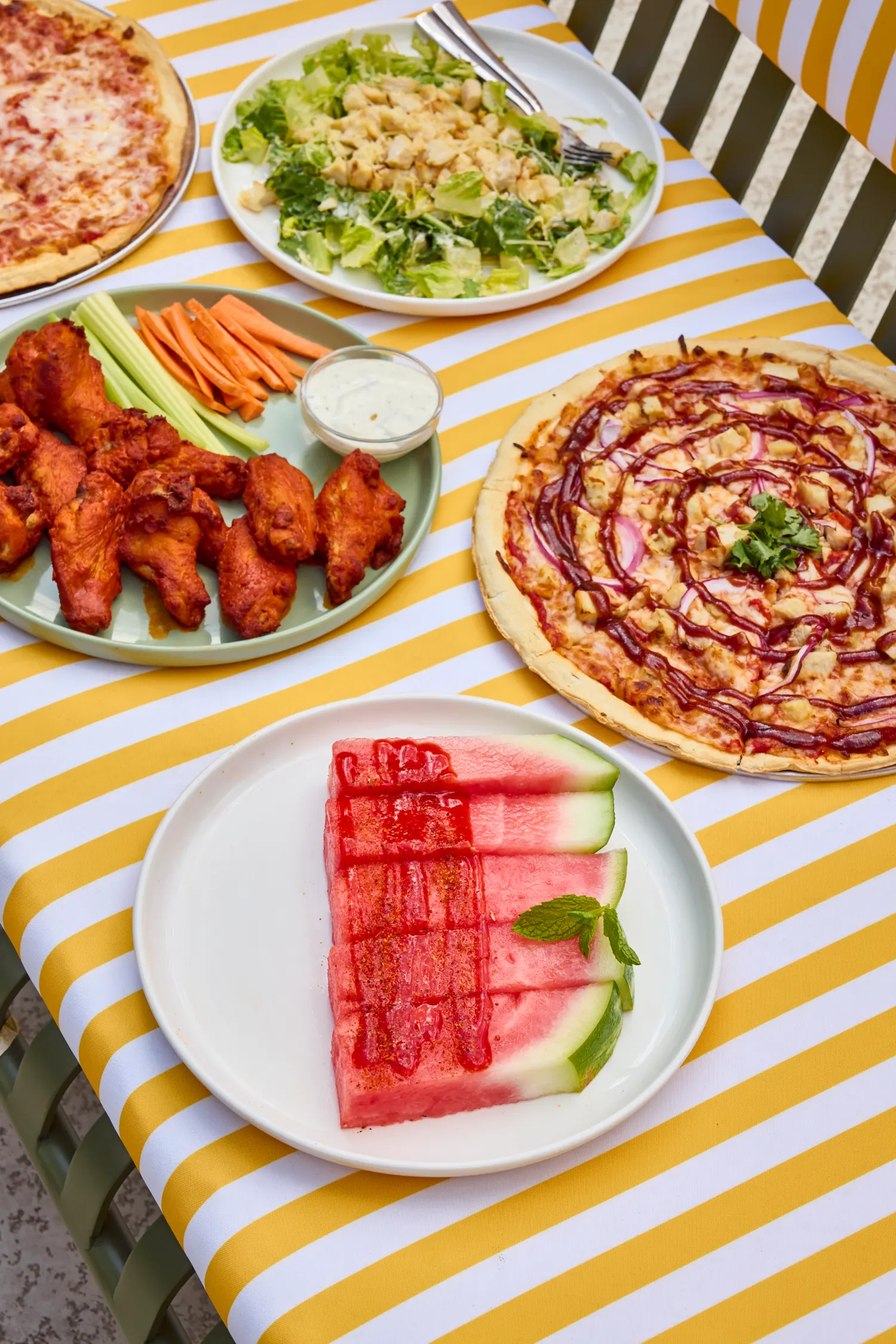 Wings, caesar, barbecue chicken pizza and chamoy watermelon on a yellow striped cloth, La Piscine